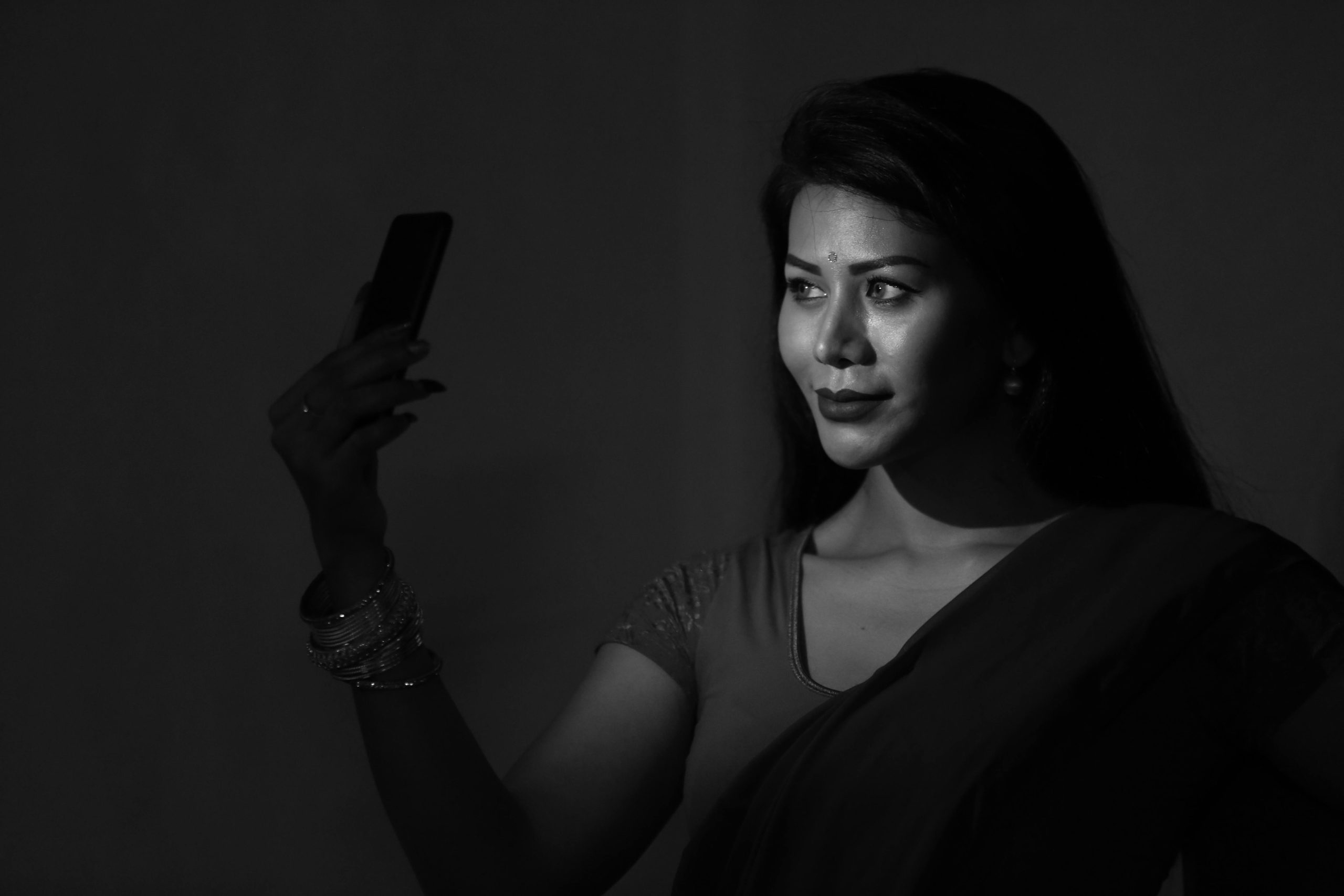 Black and white portrait of an Indian woman in a sari taking a selfie, highlighting grace and technology.