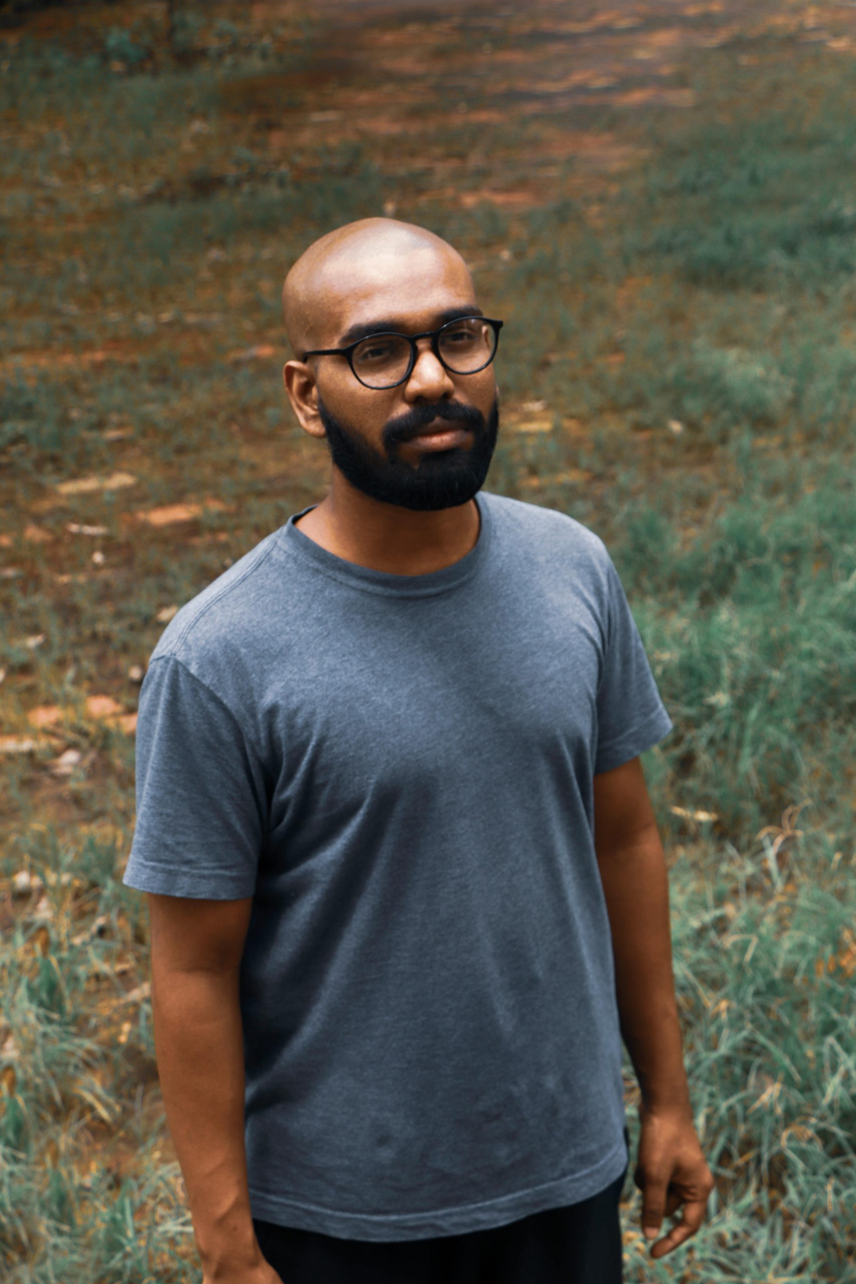 Portrait of a bald, bearded man with eyeglasses standing outdoors, wearing a gray shirt on a grassy field in India.