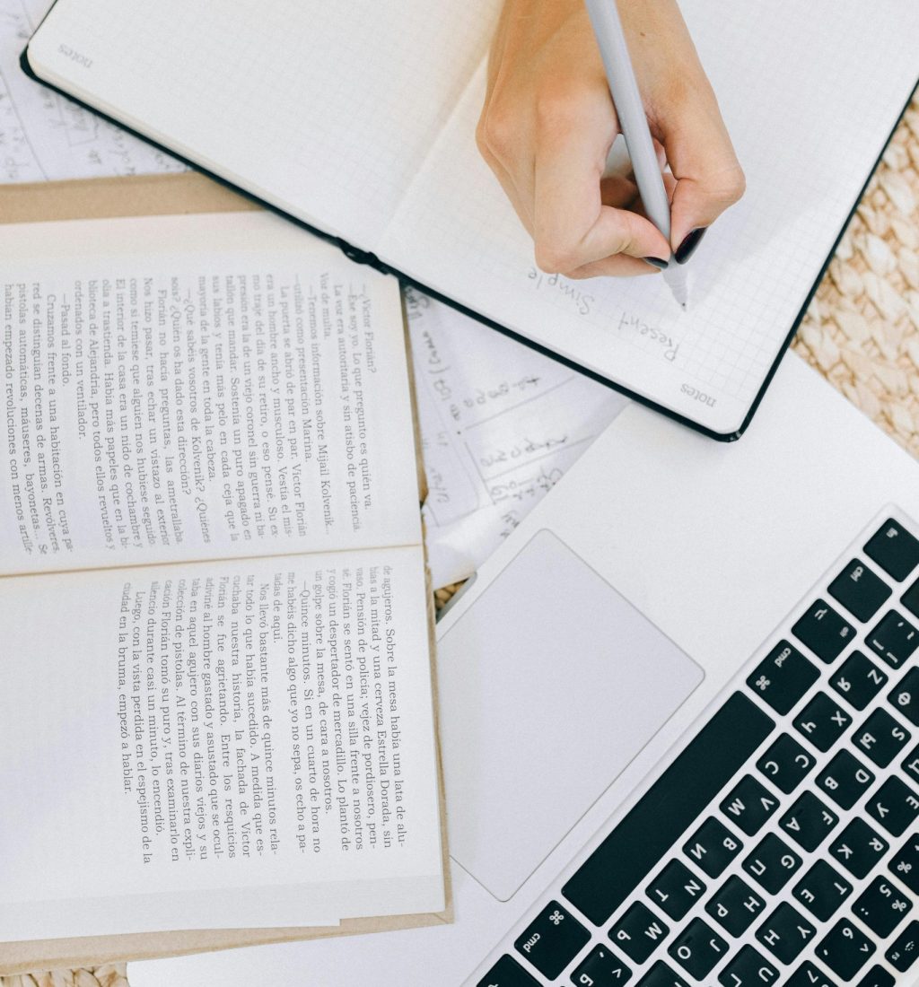 Close-up of a woman's hand writing in an open notebook on a desk with a laptop and books.